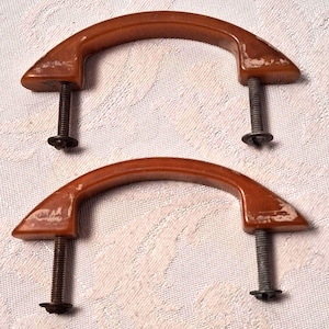 May include: Two arched, brown, semi-glossy drawer pulls with visible surface wear and paint loss. Each handle features two threaded metal bolts extending downward, complete with small circular metal washers and nuts attached at the ends.