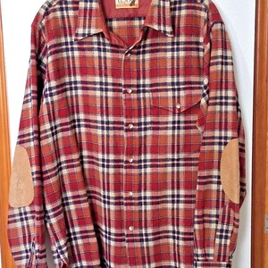 May include: A long-sleeve button-down shirt featuring a red, orange, and navy blue plaid pattern. The shirt includes a collar, a single chest pocket, and tan suede-like elbow patches. A label inside the collar displays the text LOBO.