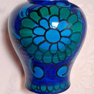 May include: A blue ceramic vase with a floral design in shades of blue and green. The vase has a rounded body and a narrow neck. The floral design is centered on the vase and is outlined in black.