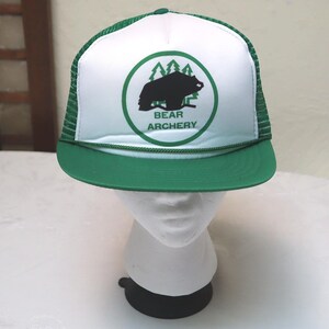 May include: A green and white trucker hat featuring a graphic of a black bear silhouette in front of green pine trees inside a circular border. The text BEAR ARCHERY is printed in green below the bear. The hat has a green brim and mesh back.