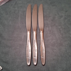 May include: Three stainless steel butter knives with a floral design on the handles. The knives are arranged vertically against a dark gray background. The blades are smooth and the handles have a decorative pattern.