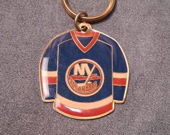 May include: A blue, red, and white metal keychain featuring the New York Islanders hockey team logo. The keychain is shaped like a hockey jersey with a gold-colored ring.