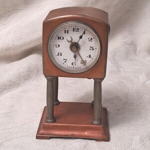 May include: A vintage copper-toned desk clock with a white clock face and black numerals. The clock has a rectangular body and is supported by two cylindrical columns. The clock face has a small second hand and the word "Wurttemberg" is printed on the clock face.