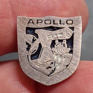 May include: A silver-toned metal pin shaped like a shield with the word APOLLO embossed at the top. The center features a dark blue recessed background with a raised silver graphic depicting a telescope and a stylized celestial element.