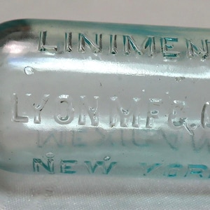 May include: A clear glass bottle with a blue tint. The bottle is labeled "Liniment" and "Lyon Mfg. & Co. New York".