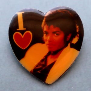 May include: Heart-shaped button featuring a portrait of Michael Jackson. The button has a black background with the words "I" and a red heart. The image of Michael Jackson is in the center, with an orange and white border.