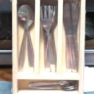 May include: A stainless steel flatware set in a wooden box. The set includes spoons, forks, and knives, neatly arranged in individual compartments. The wooden box has a natural finish, and the flatware is wrapped in clear plastic.