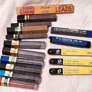 May include: A collection of various mechanical pencil lead refill containers in assorted colors and brands, including Scripto, Pentel, Sanford, and W.A. Sheaffer Pen Co. The containers feature printed text indicating lead sizes, types, and prices.