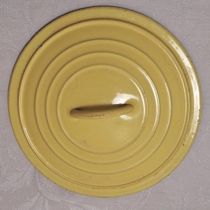 May include: A round, yellow pot lid with concentric rings and a loop handle. The lid is made of metal and has a slightly worn appearance. The handle is a lighter shade of yellow.