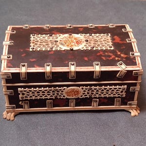 May include: A rectangular antique box with a dark brown tortoiseshell pattern. The box is trimmed with ivory-colored accents and decorative panels. The top and front feature ornate, openwork designs with floral details. The box rests on small, carved feet.