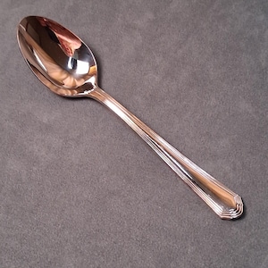 May include: A shiny, silver spoon with a rounded bowl and a long, detailed handle. The spoon reflects the surrounding environment, highlighting its polished surface. The handle features a series of parallel lines, adding to its elegant design. This utensil is suitable for serving.