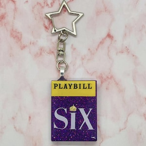 Puede incluir: Llavero de purpurina morada con un 'PLAYBILL' dorado y 'Six' con una corona en la parte superior.