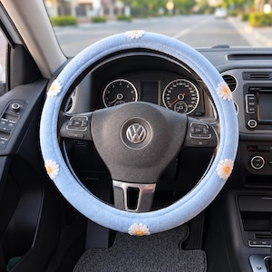 May include: A light blue steering wheel cover featuring a soft, plush texture and embroidered white daisy flowers with yellow centers spaced evenly around the circumference, installed on a black Volkswagen steering wheel with a central logo.