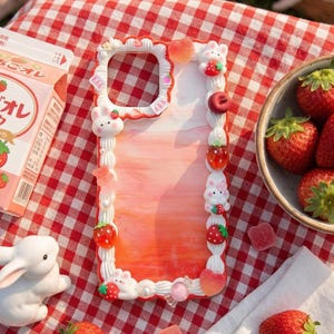 Puede incluir: Una funda decorativa para teléfono con un diseño de mármol rosa y blanco, adornada con fresas y figuras de conejos. También hay un cartón de leche de fresa y un cuenco de fresas frescas. La escena está ambientada en una manta de picnic a cuadros rojos y blancos.