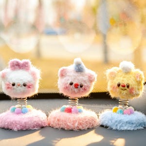 May include: Three plush, bobblehead-style car accessories. Each has a spring base and a fluffy, animal-like head with a smiling face. The heads are white with a pink bow, pink with a blue hat, and yellow with a white pom-pom. The bases are pink, pink, and blue.