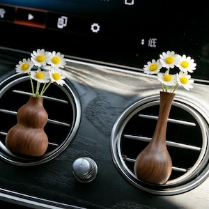 May include: Two small, wooden vases, each holding a small bouquet of white daisies with yellow centers, are displayed in a car's air vents. The vases have a natural wood finish and are set against a dark dashboard.