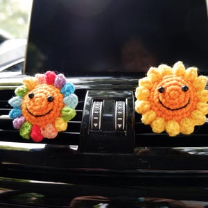 May include: Two crocheted sun-shaped car air vent decorations. One sun has a rainbow of colors around the face, and the other is yellow. Both have black eyes and a smiling mouth. The decorations are placed on a black car dashboard.