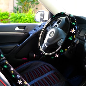 May include: Car interior featuring a black steering wheel cover and seatbelt cover, decorated with embroidered flowers in various colours. The seat cover is black with red edging. The steering wheel has the Volkswagen logo.