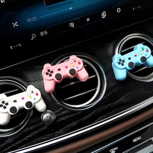 Op de afbeelding: Drie kleurrijke auto-ventilatieclips in de vorm van videogamecontrollers. Eén witte, één roze en één lichtblauwe. Elke controller heeft zwarte knoppen en kleurrijke directionele pads. De controllers zijn geplaatst op een zwart dashboard.