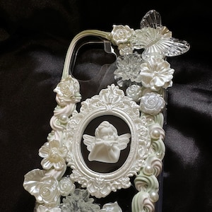 May include: A clear phone case decorated with white and pale green sculpted floral elements, small white pearls, a translucent butterfly, and a central oval frame containing a white cherub figurine. The design features intricate relief details.
