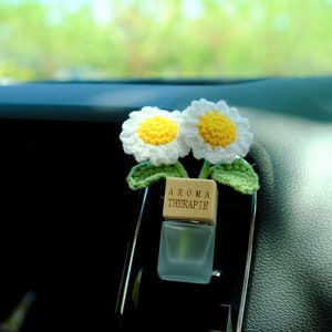 May include: A white crocheted flower car air freshener with a wooden block that says "Aroma Therapie" and a clear glass bottle.