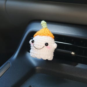 May include: A white crocheted air vent charm shaped like a smiling ghost with a yellow and green hat. The charm has black button eyes and pink cheeks. The charm is attached to a car air vent.