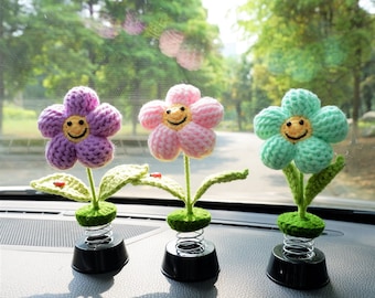 Könnte beinhalten: Drei lächelnde gehäkelte Blumen-Auto-Armaturenbrett-Dekorationen. Die Blumen sind lila, rosa und grün. Jede Blume ist an einer Feder befestigt.