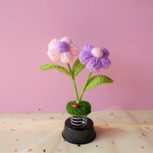 May include: A spring-loaded flower pot with two yarn flowers, one pink and one purple, with green leaves. A small red ladybug is on the green base.
