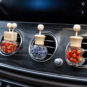 Puede incluir: Tres ambientadores de coche fijados en rejillas de ventilación circulares. El de la izquierda es un frasco de cristal cuadrado con fresas rojas y un tapón de madera con la inscripción AROMA THERAPIE. El central contiene arándanos y el de la derecha tiene forma de manzana.