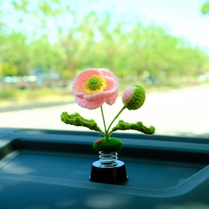 May include: A pink and yellow crocheted flower with green leaves attached to a spring-loaded base. The flower is a car decoration.