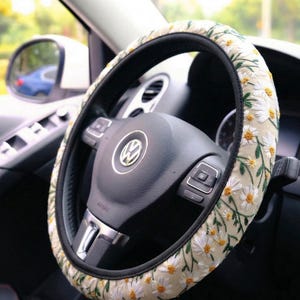 May include: A steering wheel cover featuring a cream-colored fabric base with an embroidered pattern of white daisy flowers with yellow centers and green stems, fitted onto a black Volkswagen steering wheel with a central silver logo emblem.