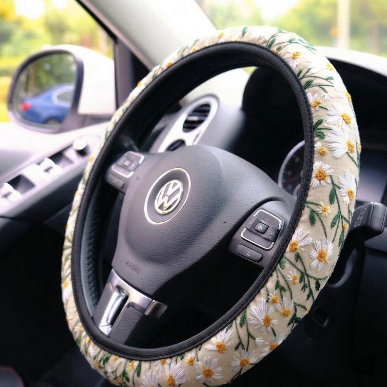 May include: A steering wheel cover featuring a cream-colored fabric base with an embroidered pattern of white daisy flowers with yellow centers and green stems, fitted onto a black Volkswagen steering wheel with a central silver logo emblem.