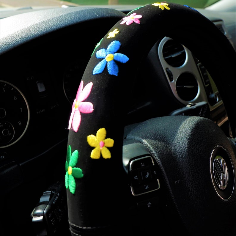May include: Black steering wheel cover decorated with embroidered flowers in pink, yellow, blue, and green. The cover is designed to fit a car's steering wheel, adding a decorative touch to the vehicle's interior.