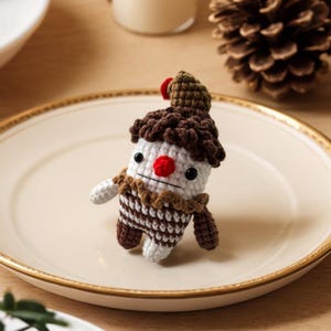 May include: A handmade crochet figure resembling a cupcake with a whimsical design. It features a white base, brown frosting, and a red cherry on top. The figure has black eyes, a red nose, and small arms and legs. It is placed on a gold-rimmed plate.