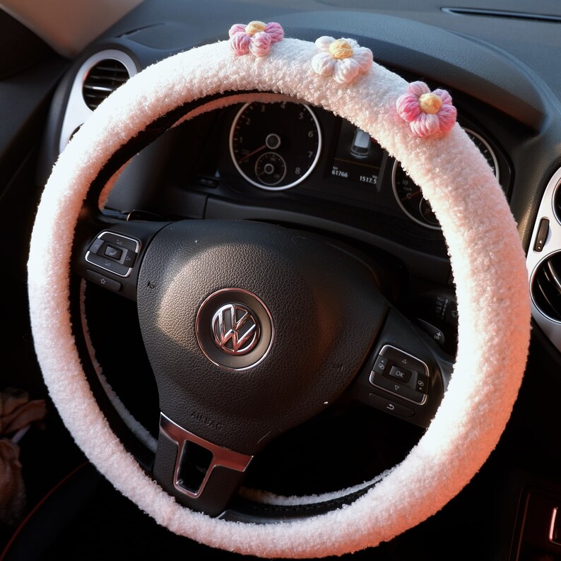 May include: White fluffy steering wheel cover with pink flower embellishments. The cover is made of soft, plush material and is designed to provide a comfortable grip on the steering wheel.