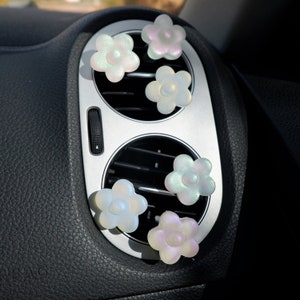 May include: Six iridescent flower-shaped car air fresheners in a car's air vent. The fresheners are in shades of white, pink, and blue.