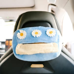 May include: A light blue car tissue holder with three embroidered daisy flowers. The tissue holder is rectangular with a slot for dispensing tissues. The holder is designed to attach to a car seat.