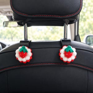 May include: Two black plastic car seat headrest hooks, each featuring a decorative, round, crocheted strawberry motif. The strawberry design consists of a red centre with white petals and a green stem, attached to the front of each hook.