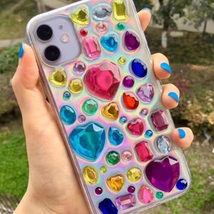 May include: Clear phone case adorned with an array of colorful, gem-like embellishments. The design includes heart, oval, and rectangular shapes in shades of pink, blue, yellow, and purple. The case has a holographic, iridescent background.