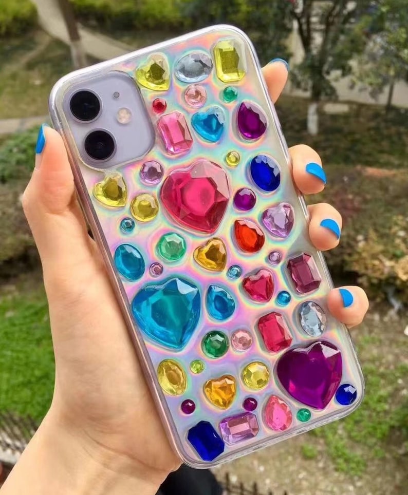 May include: Clear phone case adorned with an array of colorful, gem-like embellishments. The design includes heart, oval, and rectangular shapes in shades of pink, blue, yellow, and purple. The case has a holographic, iridescent background.