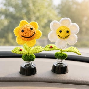 May include: Two crochet flower car decorations. One is yellow, the other is white, both with smiling faces. Each flower has green leaves and a red ladybird. They are mounted on black bases with springs.