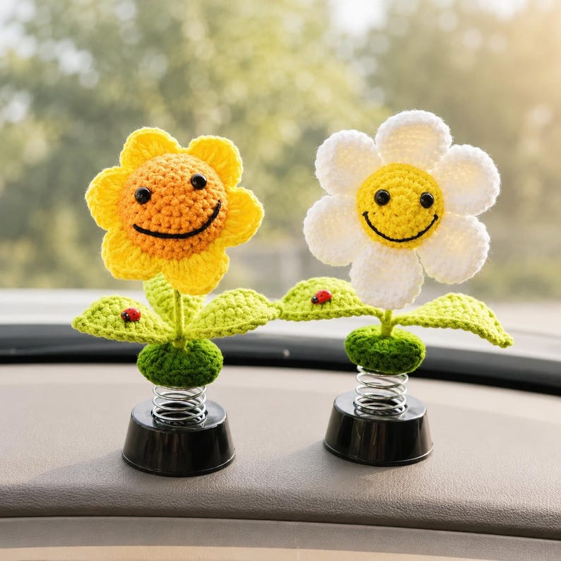 May include: Two crochet flower car decorations. One is yellow, the other is white, both with smiling faces. Each flower has green leaves and a red ladybird. They are mounted on black bases with springs.