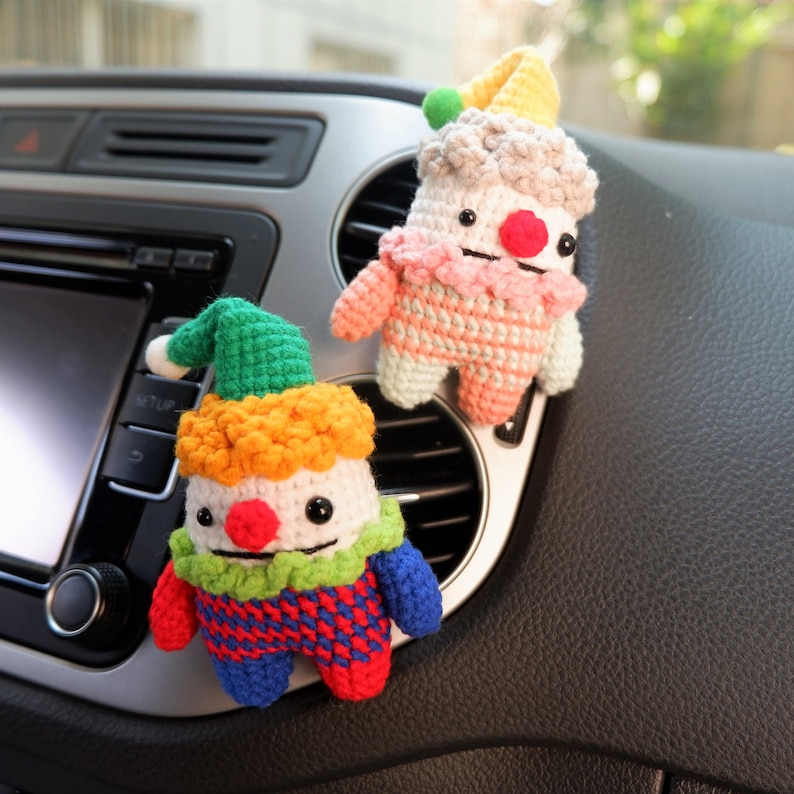 May include: Two crocheted clown dolls with colourful clothing and yarn hair hanging from the air vent of a car. The clown on the left is wearing a green hat, red and blue checked shirt, and orange hair. The clown on the right is wearing a pink and white striped shirt, a yellow hat, and white hair.