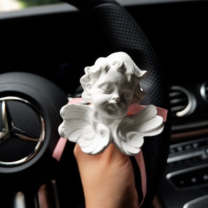 May include: White porcelain cherub figurine with wings, holding a pink ribbon. The figurine is attached to a car's steering wheel.