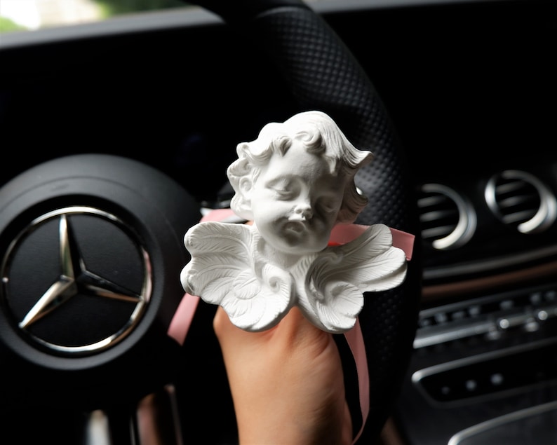 May include: White porcelain cherub figurine with wings, holding a pink ribbon. The figurine is attached to a car's steering wheel.