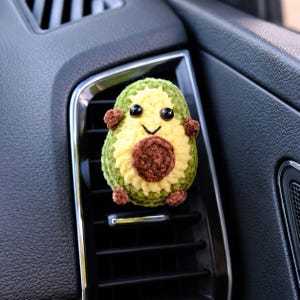 Puede incluir: Un pequeño adorno de ganchillo con forma de aguacate, con una capa exterior verde texturizada, un centro amarillo pálido y un hueso marrón oscuro. Tiene dos ojos de cuentas negras, una sonrisa negra bordada y cuatro pequeñas extremidades marrones.