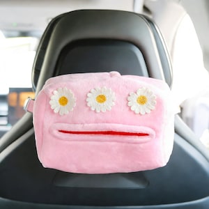 May include: A pink plush tissue box holder with three daisy flower appliqués. The tissue box holder has a red slit for tissue dispensing and a strap for hanging in a car. The holder is attached to a car seat.
