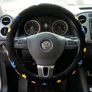 May include: Black steering wheel cover decorated with embroidered flowers in various colours. The cover is designed for a standard car steering wheel. The car's dashboard and gauges are visible in the background, with the Volkswagen logo at the centre of the steering wheel.