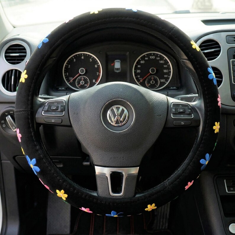 May include: Black steering wheel cover decorated with embroidered flowers in various colours. The cover is designed for a standard car steering wheel. The car's dashboard and gauges are visible in the background, with the Volkswagen logo at the centre of the steering wheel.