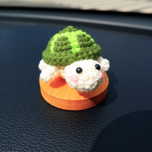 May include: A handmade crochet turtle figurine. The turtle has a green and light green shell, a white head and limbs, and black eyes. It sits on a small, round, orange wooden base. The turtle's cheeks are lightly blushed pink.